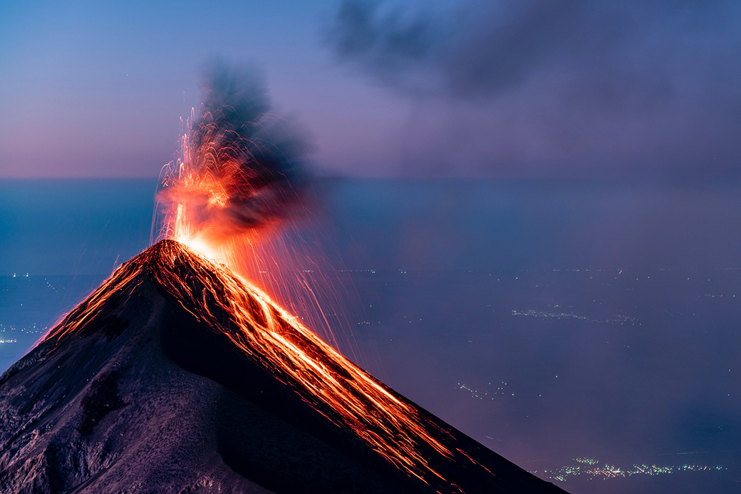 Volcanic eruption of a strato-volcano with lava running down the side, with urban settlements seen in the background.